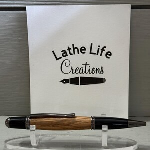 Pu&ograve; includere: Una penna artigianale con fusto in legno e dettagli neri, esposta su un supporto trasparente. La penna ha una clip e una punta argentate. Dietro la penna c'&egrave; una carta bianca con il testo "Lathe Life Creations" e un'illustrazione di pennino.