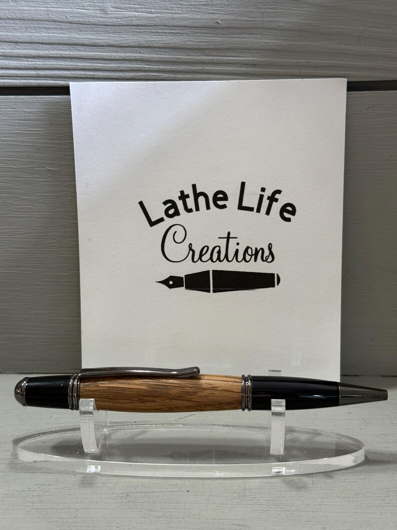 Pu&ograve; includere: Una penna artigianale con fusto in legno e dettagli neri, esposta su un supporto trasparente. La penna ha una clip e una punta argentate. Dietro la penna c'&egrave; una carta bianca con il testo "Lathe Life Creations" e un'illustrazione di pennino.