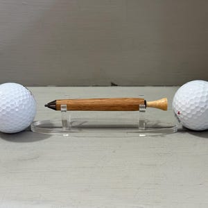 Puede incluir: Un bol&iacute;grafo de madera con acabado marr&oacute;n claro, exhibido sobre un soporte acr&iacute;lico transparente, flanqueado por dos pelotas de golf blancas. El bol&iacute;grafo tiene una punta oscura y un extremo de color claro. Las pelotas de golf tienen una superficie con hoyuelos.
