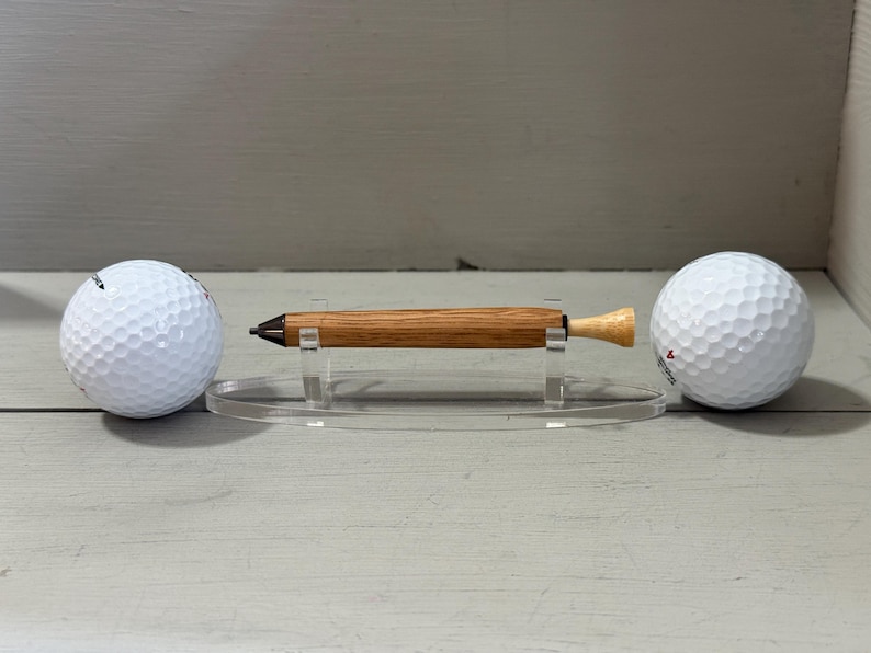 Puede incluir: Un bol&iacute;grafo de madera con acabado marr&oacute;n claro, exhibido sobre un soporte acr&iacute;lico transparente, flanqueado por dos pelotas de golf blancas. El bol&iacute;grafo tiene una punta oscura y un extremo de color claro. Las pelotas de golf tienen una superficie con hoyuelos.
