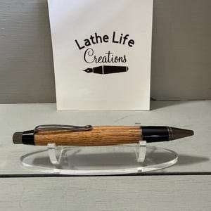 Puede incluir: Un bol&iacute;grafo de madera con un cuerpo marr&oacute;n claro, detalles negros y un clip plateado. El bol&iacute;grafo se exhibe sobre un soporte de acr&iacute;lico transparente frente a una tarjeta blanca con el texto "Lathe Life Creations".
