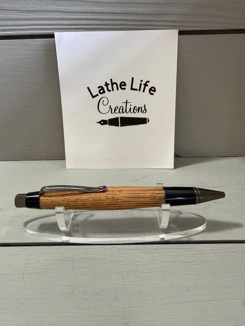 Puede incluir: Un bol&iacute;grafo de madera con un cuerpo marr&oacute;n claro, detalles negros y un clip plateado. El bol&iacute;grafo se exhibe sobre un soporte de acr&iacute;lico transparente frente a una tarjeta blanca con el texto "Lathe Life Creations".