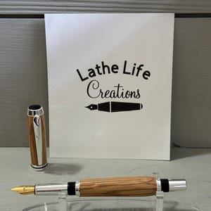 May include: A handcrafted fountain pen with a wooden barrel and silver accents, displayed on a clear acrylic stand. The pen features a gold nib and a matching cap. The background includes a white card with the text "Lathe Life Creations".