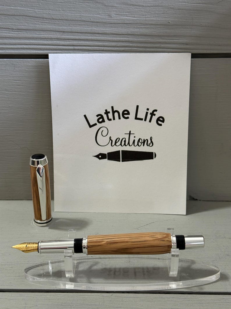 May include: A handcrafted fountain pen with a wooden barrel and silver accents, displayed on a clear acrylic stand. The pen features a gold nib and a matching cap. The background includes a white card with the text "Lathe Life Creations".