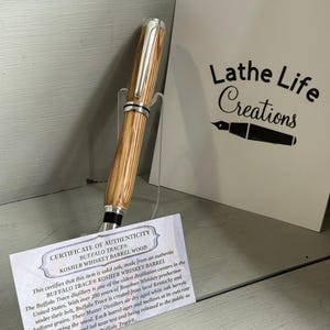 May include: A wooden pen with silver accents, displayed on a clear stand. The pen is made from Buffalo Trace Kosher Whiskey Barrel wood. A card with the text "Lathe Life Creations" and a certificate of authenticity are in the background.