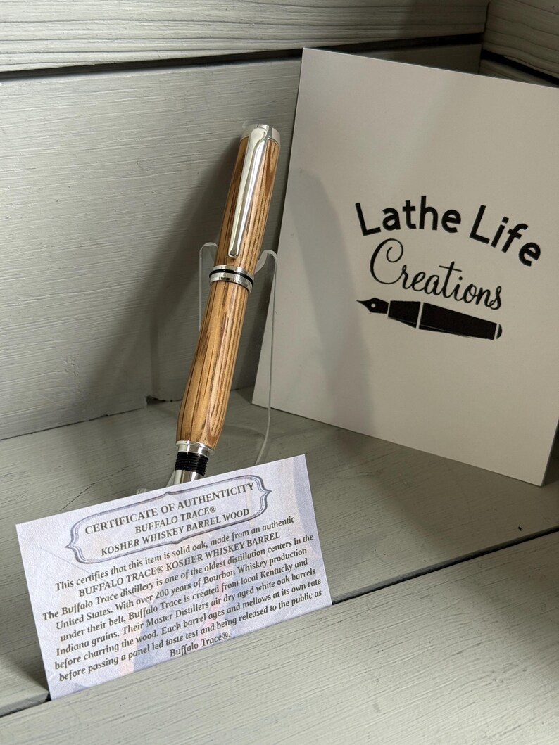 May include: A wooden pen with silver accents, displayed on a clear stand. The pen is made from Buffalo Trace Kosher Whiskey Barrel wood. A card with the text "Lathe Life Creations" and a certificate of authenticity are in the background.