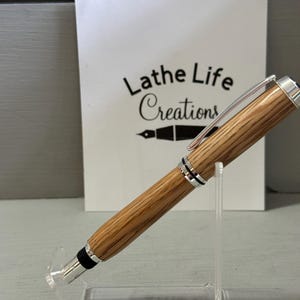 May include: A wooden pen with silver accents and a black tip is displayed on a clear acrylic stand. The pen has a light brown, striped pattern. Behind the pen is a white card with the text "Lathe Life Creations".