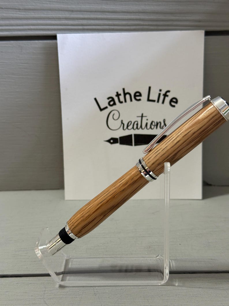 May include: A wooden pen with silver accents and a black tip is displayed on a clear acrylic stand. The pen has a light brown, striped pattern. Behind the pen is a white card with the text "Lathe Life Creations".
