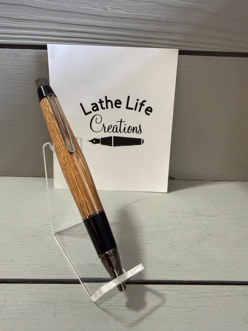Puede incluir: Un bol&iacute;grafo de madera con acabado marr&oacute;n claro y detalles en negro, expuesto sobre un soporte de acr&iacute;lico transparente. El bol&iacute;grafo tiene un clip y una punta plateados. Una tarjeta blanca detr&aacute;s del bol&iacute;grafo dice "Lathe Life Creations".
