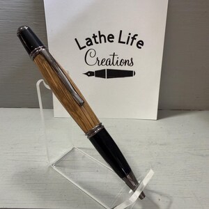 Pu&ograve; includere: Una penna in legno fatta a mano con dettagli neri e finiture color argento &egrave; esposta su un supporto in acrilico trasparente. La penna ha un fusto in legno marrone chiaro. Una carta bianca dietro la penna recita "Lathe Life Creations".