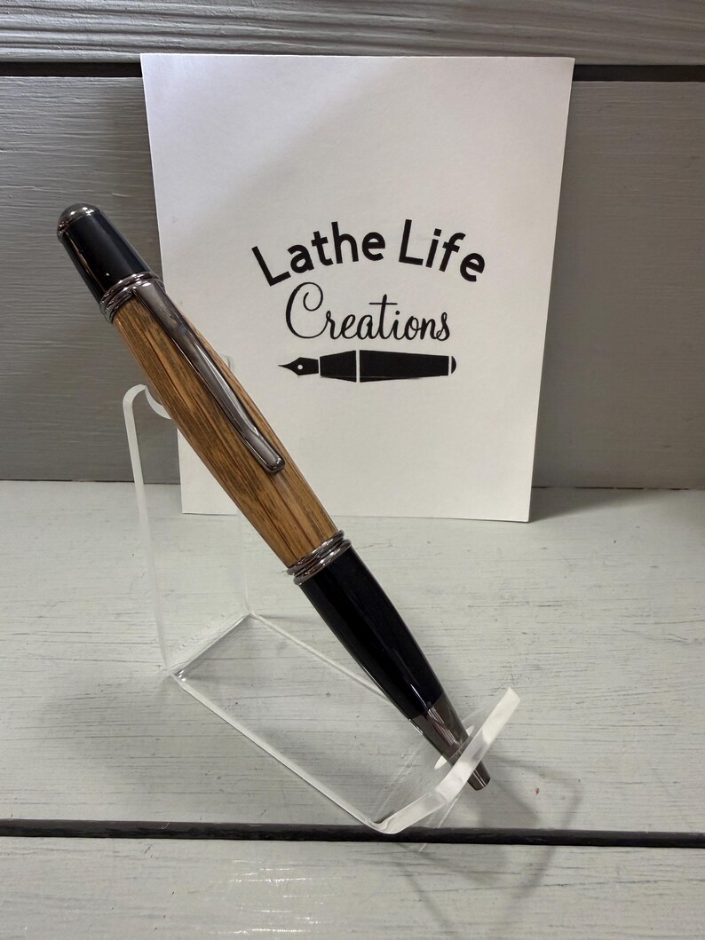 Pu&ograve; includere: Una penna in legno fatta a mano con dettagli neri e finiture color argento &egrave; esposta su un supporto in acrilico trasparente. La penna ha un fusto in legno marrone chiaro. Una carta bianca dietro la penna recita "Lathe Life Creations".