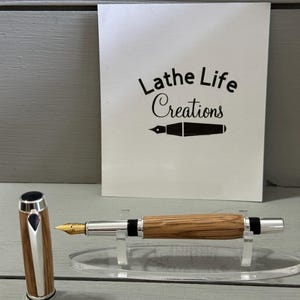 May include: A wooden fountain pen with a silver-colored tip and accents, and a gold-colored nib. The pen is displayed on a clear acrylic stand. The pen cap is next to the pen. A white card with the text "Lathe Life Creations" is in the background.