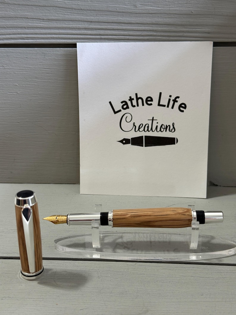 May include: A wooden fountain pen with a silver-colored tip and accents, and a gold-colored nib. The pen is displayed on a clear acrylic stand. The pen cap is next to the pen. A white card with the text "Lathe Life Creations" is in the background.