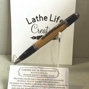 Pu&ograve; includere: Una penna in legno con dettagli neri e finiture argentate, esposta su un supporto trasparente. La penna &egrave; realizzata in legno di botte di whisky Buffalo Trace Kosher. Un certificato di autenticit&agrave; &egrave; visibile con il testo "Lathe Life Creations."
