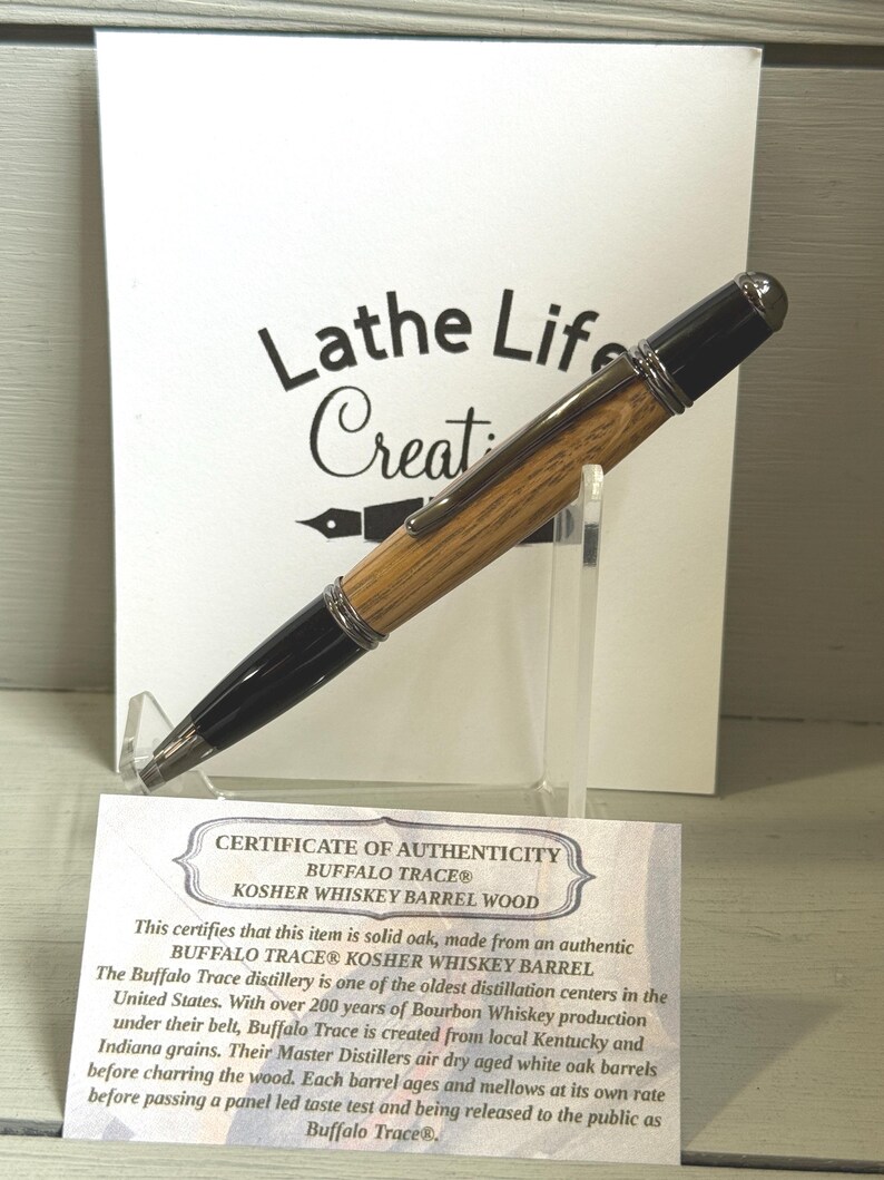 Pu&ograve; includere: Una penna in legno con dettagli neri e finiture argentate, esposta su un supporto trasparente. La penna &egrave; realizzata in legno di botte di whisky Buffalo Trace Kosher. Un certificato di autenticit&agrave; &egrave; visibile con il testo "Lathe Life Creations."