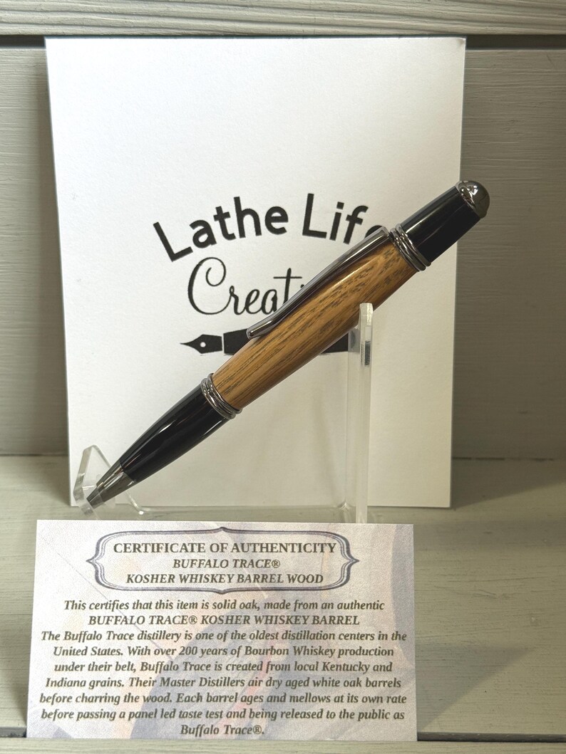 Pu&ograve; includere: Una penna in legno con dettagli neri, presentata su un supporto trasparente. La penna &egrave; realizzata in legno di botte di whisky Buffalo Trace Kosher. Un certificato di autenticit&agrave; e un cartello con il testo "Lathe Life Creat" sono sullo sfondo.