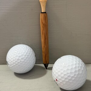 Puede incluir: Imagen de dos pelotas de golf blancas y un bol&iacute;grafo de golf de madera con una parte superior en forma de tee. El bol&iacute;grafo tiene una punta oscura y un cuerpo de madera marr&oacute;n claro. Las pelotas de golf tienen una superficie con hoyuelos. El bol&iacute;grafo mide unos 15 cm.