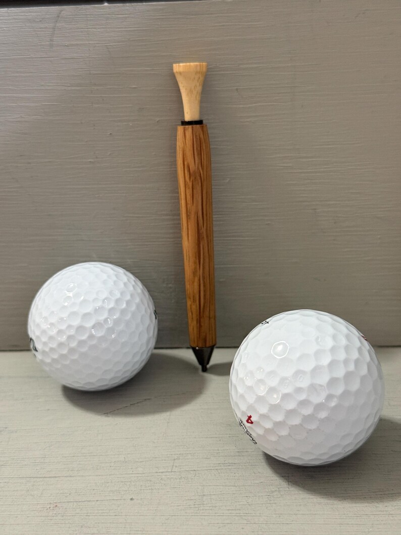 Puede incluir: Imagen de dos pelotas de golf blancas y un bol&iacute;grafo de golf de madera con una parte superior en forma de tee. El bol&iacute;grafo tiene una punta oscura y un cuerpo de madera marr&oacute;n claro. Las pelotas de golf tienen una superficie con hoyuelos. El bol&iacute;grafo mide unos 15 cm.