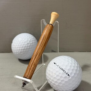 Puede incluir: Un bol&iacute;grafo con tem&aacute;tica de golf con un cuerpo de madera y una parte superior de color claro, exhibido en un soporte transparente junto a dos pelotas de golf blancas. Una pelota de golf tiene impreso "PRO SERIES 2". El bol&iacute;grafo est&aacute; dise&ntilde;ado para parecerse a un tee de golf.