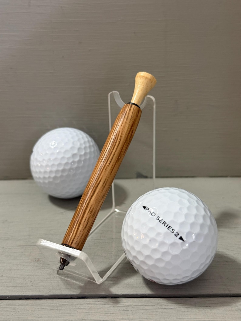 Puede incluir: Un bol&iacute;grafo con tem&aacute;tica de golf con un cuerpo de madera y una parte superior de color claro, exhibido en un soporte transparente junto a dos pelotas de golf blancas. Una pelota de golf tiene impreso "PRO SERIES 2". El bol&iacute;grafo est&aacute; dise&ntilde;ado para parecerse a un tee de golf.