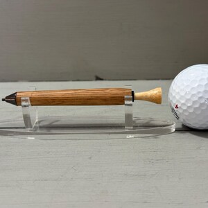 Puede incluir: Un bol&iacute;grafo de madera con punta oscura y extremo de color claro, exhibido sobre un soporte transparente junto a una pelota de golf blanca. El bol&iacute;grafo tiene un acabado de veta de madera natural.