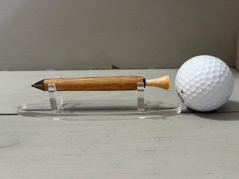 Puede incluir: Un bol&iacute;grafo de madera con punta oscura y extremo de color claro, exhibido sobre un soporte transparente junto a una pelota de golf blanca. El bol&iacute;grafo tiene un acabado de veta de madera natural.