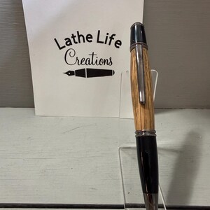 Pu&ograve; includere: Una penna artigianale con fusto in legno e dettagli neri, esposta su un supporto in acrilico trasparente. La penna ha una clip e una punta argentate. Una scheda bianca dietro la penna recita "Lathe Life Creations".