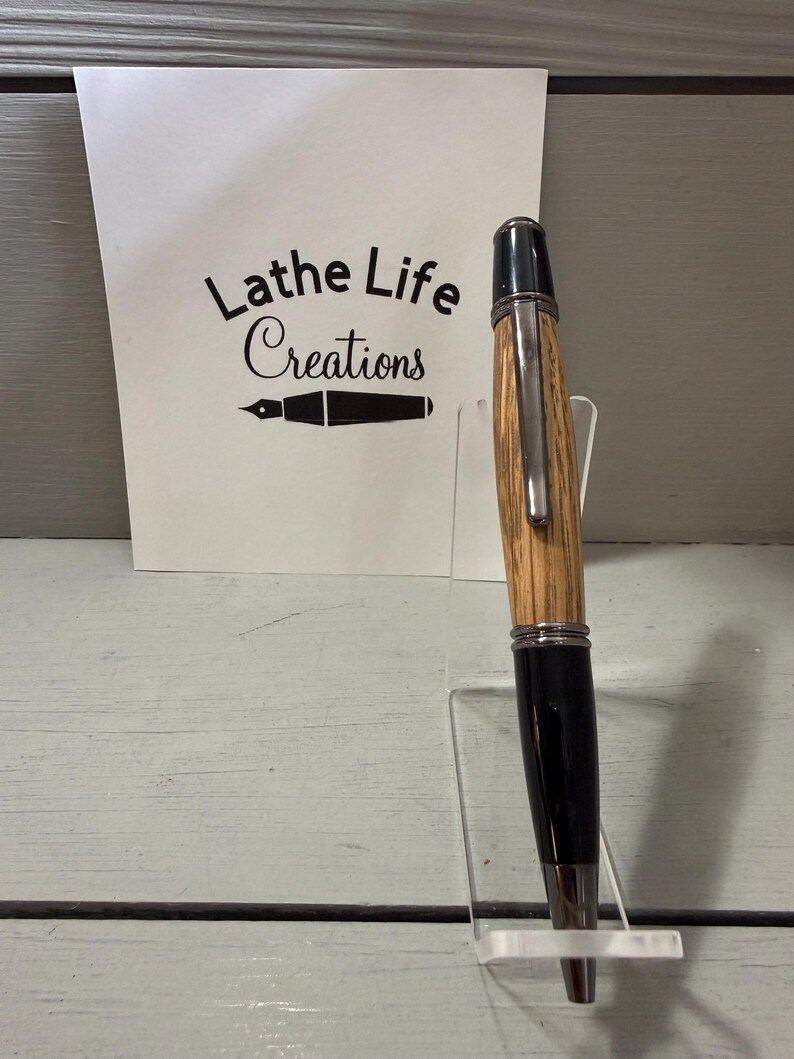 Pu&ograve; includere: Una penna artigianale con fusto in legno e dettagli neri, esposta su un supporto in acrilico trasparente. La penna ha una clip e una punta argentate. Una scheda bianca dietro la penna recita "Lathe Life Creations".