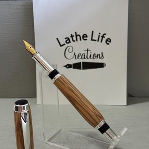 May include: A handcrafted fountain pen with a wooden barrel and silver accents. The pen features a gold nib and a matching cap. The pen is displayed on a clear acrylic stand. The text "Lathe Life Creations" is visible in the background.