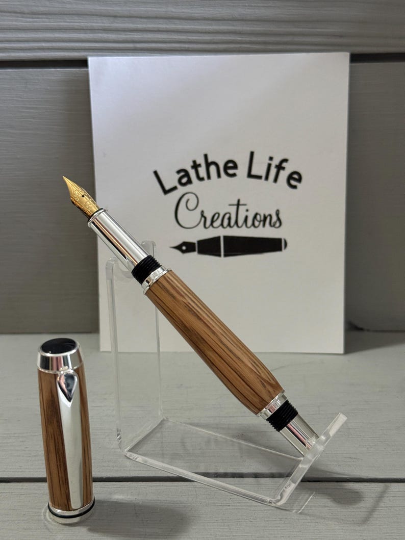 May include: A handcrafted fountain pen with a wooden barrel and silver accents. The pen features a gold nib and a matching cap. The pen is displayed on a clear acrylic stand. The text "Lathe Life Creations" is visible in the background.