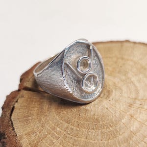 Puede incluir: Un anillo plateado con una banda texturizada y una cara circular. La cara presenta dos piedras redondas y claras engastadas en un diseño estilizado. El anillo se exhibe sobre una rodaja de madera natural.