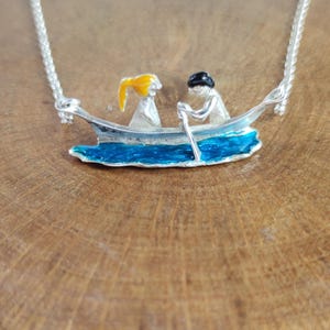 May include: Silver-toned necklace featuring a miniature boat with two figures. One figure has yellow hair, the other has black hair, and is holding an oar. The boat is painted with blue water and sits on a wooden surface.