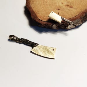 May include: A gold-colored cleaver pendant with a black handle. The pendant is on a white surface.