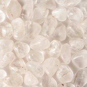 May include: A collection of tumbled clear quartz stones. The stones are smooth and polished, with a translucent appearance. They are arranged in a random pattern on a white background.