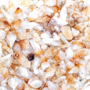 Puede incluir: Una colección de pequeños cristales de cuarzo transparentes con un tono dorado. Los cristales están dispersos sobre una superficie blanca y son de diversas formas y tamaños.