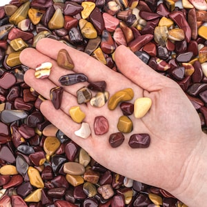 May include: A hand holding a variety of small, smooth, polished stones in shades of red, yellow, and brown. The stones are scattered on a surface.