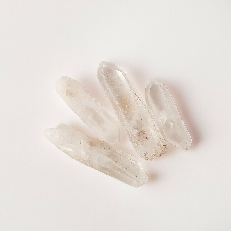 Puede incluir: Cuatro puntas de cristal de cuarzo transparente con una textura rugosa y natural. Los cristales est&aacute;n dispuestos en forma de abanico sobre un fondo blanco.
