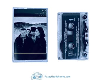 May include: A clear cassette tape of the U2 album "The Joshua Tree" with a black and white photo of the band. The cassette is next to its case, which has the album title and band name.