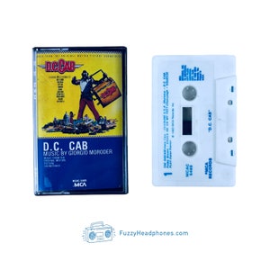 May include: A white cassette tape with blue text that reads "MCA RECORDS" and "D.C. CAB". The cassette tape is next to a black cassette case with a blue and white label that reads "D.C. CAB" and "MUSIC BY GIORGIO MORODER".