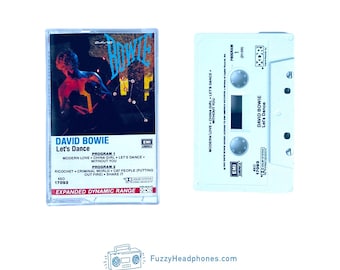 Pode incluir: Uma cassete de David Bowie Let's Dance numa caixa de plástico transparente. A capa apresenta uma imagem escura com texto azul e uma lista de faixas. A cassete branca exibe o nome do artista, o título do álbum e detalhes do programa.