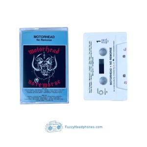 May include: A Motorhead No Remorse cassette tape with a light blue J-card featuring a black and white skull graphic and track listings. The white cassette shell displays the album title, track list, and record label information in black text.