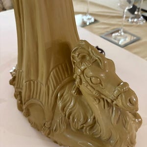 May include: A tan-colored decorative table base featuring a detailed camel sculpture. The base has a draped design and is made of a glossy material. The camel is in a seated position with intricate facial features.