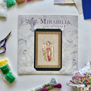 Può includere: Modello di punto croce di Mirabilia con una donna in un abito fluido con un motivo floreale. Il modello è incorniciato e circondato da rose bianche.