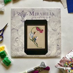 Op de afbeelding: Een kruissteekpatroon met een fee die op een bloem zit met een vlinder op haar vleugel. Het patroon is getiteld "Mirabilia cross-stitch".