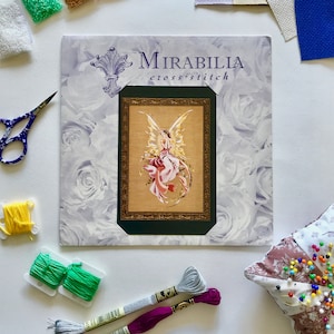 Könnte beinhalten: Ein Mirabilia Kreuzstich-Set mit einem gerahmten Feen-Design in Rosa und Gold. Das Set enthält Garnrollen in Gelb, Grün, Grau und Lila, eine Schere und ein Nadelkissen. Der Hintergrund weist ein Blumenmuster auf.