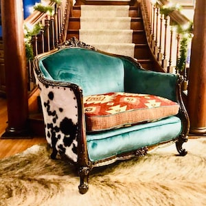 May include: An ornate teal armchair with a cowhide back and a patterned cushion. The chair has dark wood trim and sits on a cowhide rug. The background features a staircase and wooden columns, creating a vintage aesthetic.