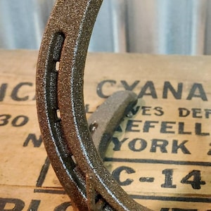 May include: A rustic brown horseshoe with a rough, textured surface. The horseshoe is lying on a wooden surface with the text "CYANAMID" and "C-14" visible.