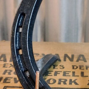 May include: A black horseshoe with a copper hook attached to the bottom. The horseshoe is sitting on a brown surface with text that reads "CANADA" and "EFELL WORK".