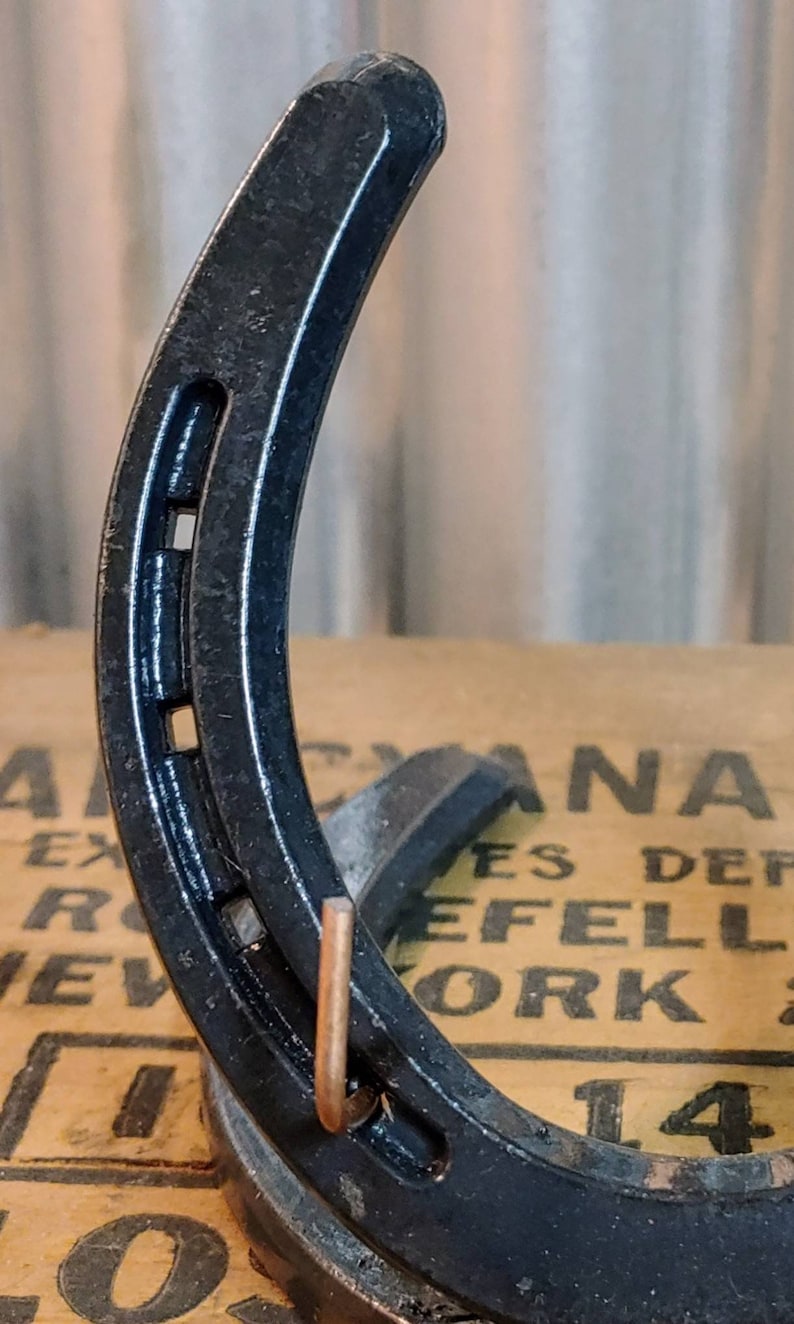 May include: A black horseshoe with a copper hook attached to the bottom. The horseshoe is sitting on a brown surface with text that reads "CANADA" and "EFELL WORK".