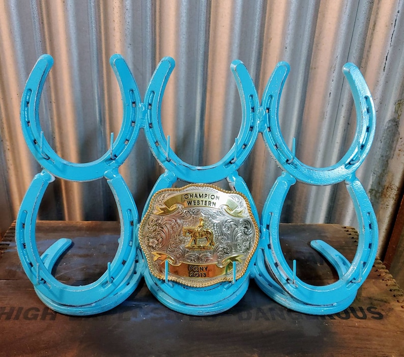 May include: A turquoise blue horseshoe rack with six horseshoes, holding a silver and gold belt buckle with the text "Champion Western" and "BQNY 2013".